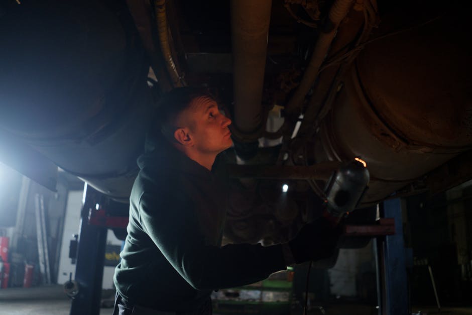 Auto mechanic inspects vehicle underbody in a garage setting with dim lighting.